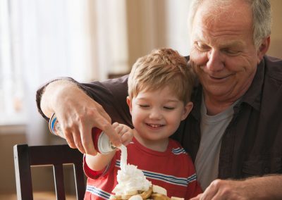 Senior man helping his grandson put large amounts of whipped cream on a stack of waffles